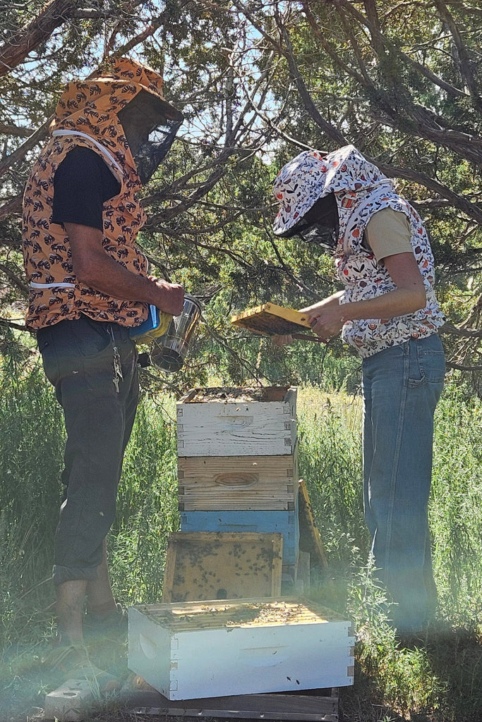 Person in beekeeping outfit standing outdoors with trees and an open beehive.